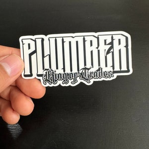May include: White sticker with black text that reads "PLUMBER" in a gothic font. Below the word "PLUMBER" is the text "King of Trades" in a smaller gothic font.