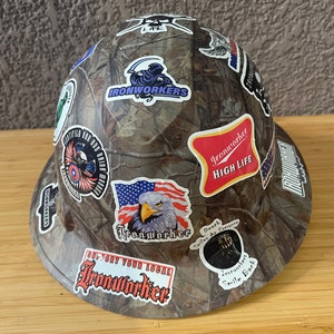 May include: A camouflage hard hat with various stickers, including a large "Ironworker" sticker with a grim reaper design, a "Ironworker High Life" sticker, and a "Support Your Local Ironworker" sticker.