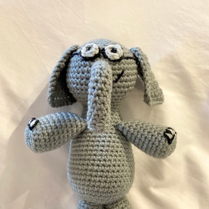 May include: A gray crocheted elephant toy wearing black framed glasses with white lenses. The elephant has a black stitched smile and black eyes.