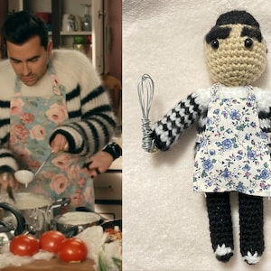 May include: A crocheted doll wearing a black and white striped sweater, a floral apron, and black pants. The doll is holding a silver whisk in its right hand.