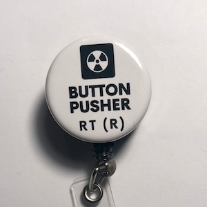 May include: White button with a black radiation symbol and the text "BUTTON PUSHER RT (R)" attached to a clear retractable badge holder.
