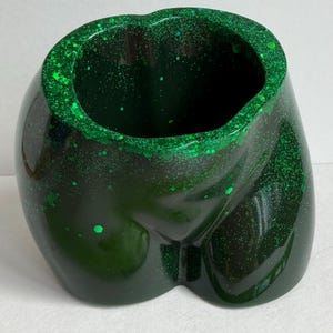 May include: A dark green, hollow, torso-shaped container crafted from a glossy material embedded with fine and chunky green glitter particles. The object features a wide, open top rim and a smooth, curved exterior surface with a deep green hue.
