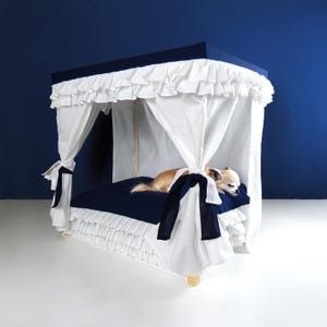 May include: A small dog rests on a navy blue pet bed with a white canopy. The bed has a ruffled skirt and a navy blue top. The canopy is trimmed with white ruffles and tied with navy blue ribbons.