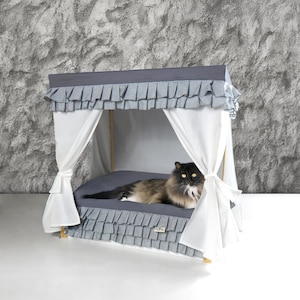 Puede incluir: Una cama para gatos gris y blanca con dosel. La cama tiene un ribete con volantes gris y blancos y un dosel blanco. Un gato gris y blanco está tumbado en la cama.
