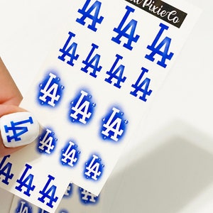 May include: A sheet of white sticker paper with multiple blue and white LA Dodgers logos. The logos are in various sizes and styles, including a spray paint effect. The text "Wicked Pixie Co" is in the top right corner of the sheet.