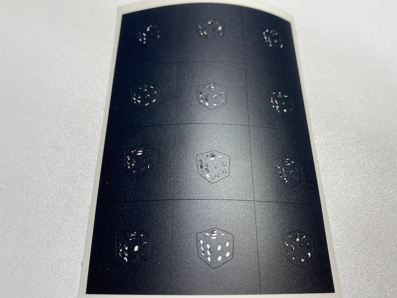 Pode incluir: Uma folha de autocolantes com uma grelha de desenhos de dados. Cada dado &eacute; retratado numa orienta&ccedil;&atilde;o diferente, com pontos brancos sobre um fundo cinzento escuro. Os autocolantes est&atilde;o sobre um suporte branco.