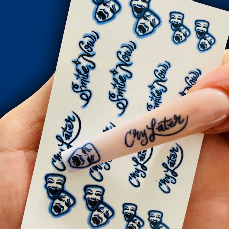 May include: A sheet of nail art stickers with a white background and blue designs. The stickers feature two different designs: a pair of theatrical masks and the phrase "Smile Now". A fingernail with a light pink polish is shown with a sticker that reads "Cry Later".