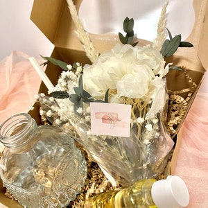 May include: A gift box filled with brown paper shreds, a clear glass bottle with a decorative design, a bottle of yellow liquid with a white cap, and a bouquet of dried white flowers with green leaves. The box is open and the items are visible.