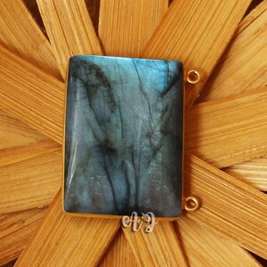 Puede incluir: Un colgante rectangular con un marco dorado y una piedra azul-verdosa. La piedra tiene un aspecto veteado con vetas oscuras. El colgante está sobre un fondo tejido de color marrón claro. Pequeños bucles dorados están unidos al colgante.