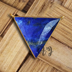 Può includere: Un ciondolo dorato con una pietra preziosa di lapislazzuli triangolare di colore blu scuro e con sfumature dorate. Il ciondolo è su una superficie di legno.