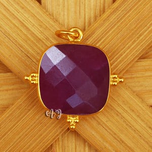 May include: A square, faceted, deep purple gemstone pendant with a gold-colored frame and bail. The pendant is set against a woven, light brown background. The gold frame has decorative accents on each side.