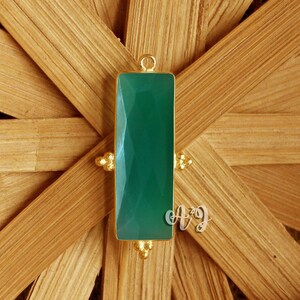 Puede incluir: Un colgante rectangular de piedra preciosa verde con una superficie facetada, engastado en un marco dorado. El colgante tiene un pequeño lazo en la parte superior para colgar y detalles dorados decorativos en la parte inferior. El fondo es un material tejido marrón claro.