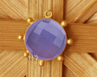 May include: A gold-colored pendant with a round, faceted, lavender-colored gemstone. The pendant has a loop at the top and is surrounded by small gold-colored spheres. The pendant is set against a wooden background.