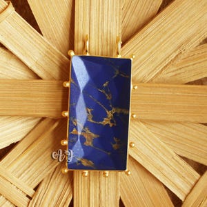 May include: A rectangular pendant with a deep blue stone and gold accents. The stone has a marbled appearance with gold veining. The pendant is set in a gold-colored frame and is displayed on a woven background.