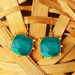 May include: A pair of gold-toned earrings with square, teal-colored stone pendants. The stones have a marbled appearance and are framed by a gold border with small, round accents. The earrings hang from gold hooks.