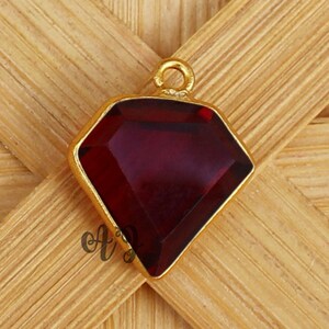 May include: A gold-toned pendant with a faceted, deep red gemstone in a diamond shape. The pendant has a small loop at the top for a chain. The background is a light brown, woven wood pattern.