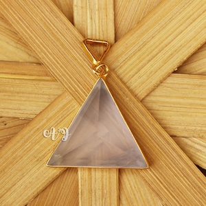 May include: A pale pink triangle pendant with a gold-tone setting. The pendant is on a wooden background.