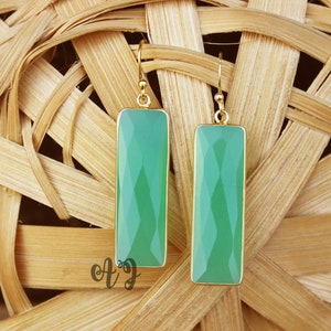 May include: A pair of gold earrings with rectangular green gemstone drops. The earrings are hanging from a woven wicker background.