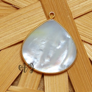 May include: A white mother of pearl teardrop pendant with a gold bail. The pendant is approximately 1.5 inches long and 1 inch wide.