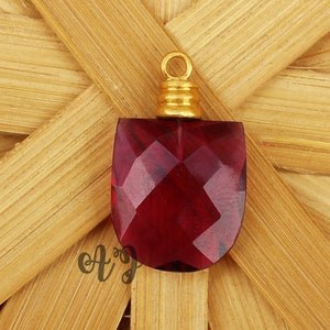 May include: A deep red faceted glass pendant with a gold-toned metal bail. The pendant is shaped like a shield and has a faceted design.