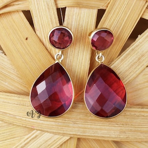 Könnte beinhalten: Ein Paar goldfarbene Ohrhänger mit facettierten, tiefroten Edelsteinen. Jeder Ohrring hat einen runden Stein oben und einen tropfenförmigen Stein unten. Die Ohrringe sind vor einem gewebten, hellbraunen Hintergrund platziert.