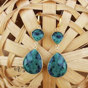 May include: A pair of gold earrings with green gemstone drops. The earrings feature two teardrop-shaped stones, one larger than the other, with a mottled green and black pattern.