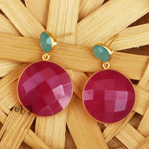 Op de afbeelding: Een paar oorhangers met een ronde, gefacetteerde, roze edelsteen en een kleinere, ronde, groene edelsteen. De oorhangers zijn gemaakt van goudkleurig metaal.