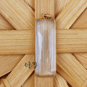 May include: A clear rectangular pendant with a gold-coloured metal frame. The pendant is on a woven wood background.