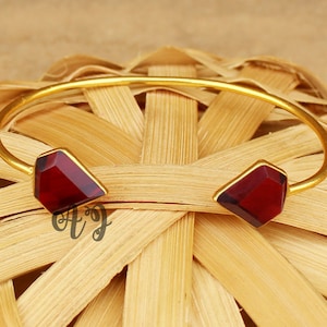 May include: Gold bangle bracelet with two red gemstone accents. The bracelet is open and has a simple design.