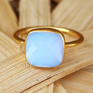 Può includere: Un anello d'oro con una pietra preziosa quadrata di colore blu chiaro. L'anello è posto su uno sfondo di bambù intrecciato.