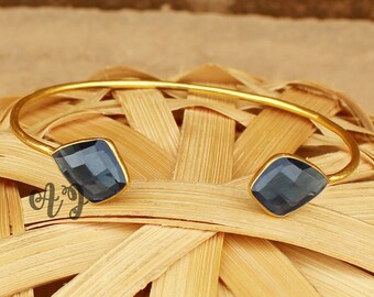 May include: Gold bangle bracelet featuring two dark blue square-shaped gemstones. The bracelet is open and has a simple design.