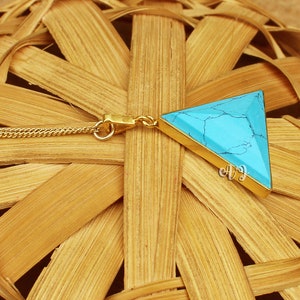 May include: A gold-colored chain necklace with a triangular pendant. The pendant features a turquoise stone set in a gold-colored frame. The necklace is displayed on a woven, light brown surface.
