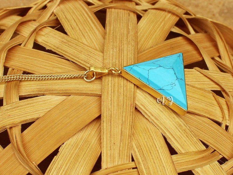 May include: A gold-colored chain necklace with a triangular pendant. The pendant features a turquoise stone set in a gold-colored frame. The necklace is displayed on a woven, light brown surface.