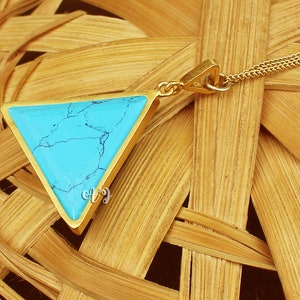 May include: A gold-colored necklace featuring a triangular pendant with a vibrant turquoise stone. The stone has a marbled appearance with dark blue veining. The pendant is suspended from a delicate gold chain, creating a simple yet elegant design.