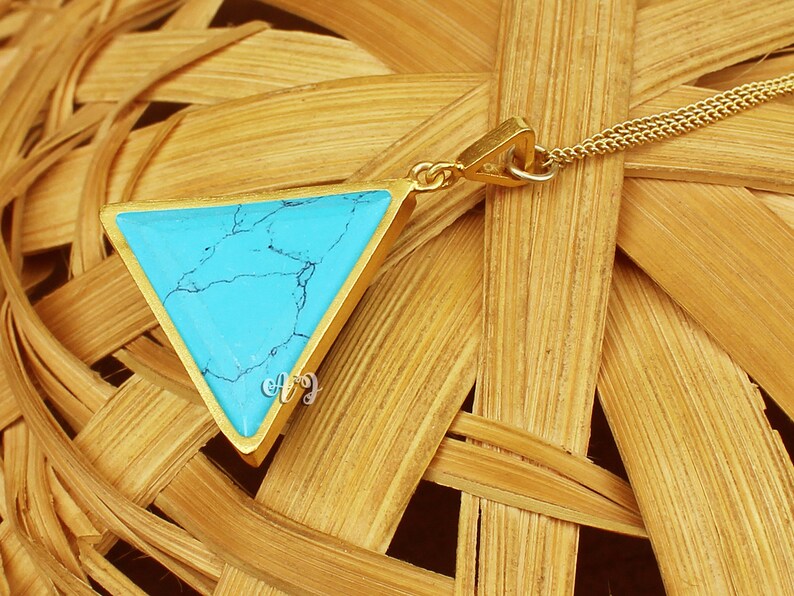 May include: A gold-colored necklace featuring a triangular pendant with a vibrant turquoise stone. The stone has a marbled appearance with dark blue veining. The pendant is suspended from a delicate gold chain, creating a simple yet elegant design.