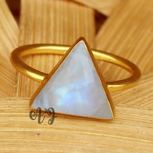 May include: Gold ring featuring a triangular moonstone gemstone. The ring is displayed on a wooden surface.