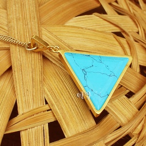 May include: A gold-colored pendant necklace featuring a turquoise triangle. The turquoise stone has dark blue veining. The pendant is suspended from a delicate gold chain. The necklace is displayed on a woven, light brown surface.