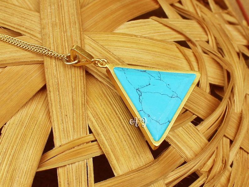 May include: A gold-colored pendant necklace featuring a turquoise triangle. The turquoise stone has dark blue veining. The pendant is suspended from a delicate gold chain. The necklace is displayed on a woven, light brown surface.