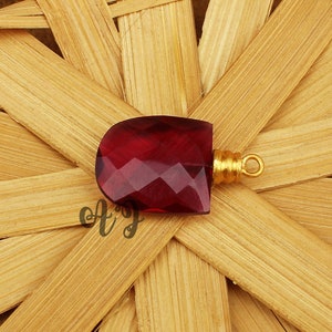 May include: A deep red faceted gemstone pendant with a gold-toned bail. The pendant is on a woven bamboo background.