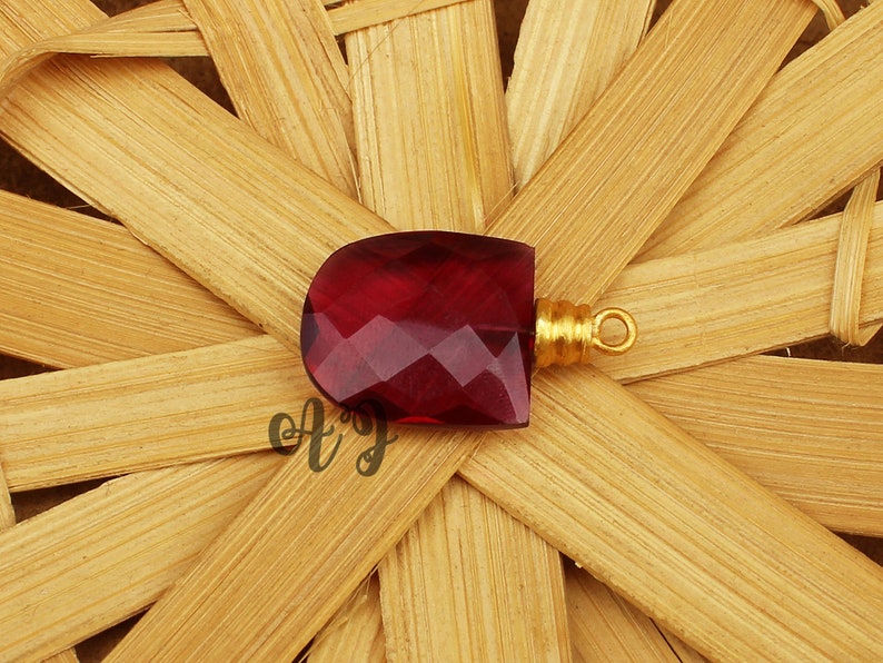 May include: A deep red faceted gemstone pendant with a gold-toned bail. The pendant is on a woven bamboo background.