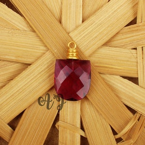May include: A deep red faceted glass pendant with a gold-toned metal bail. The pendant is on a woven straw background.