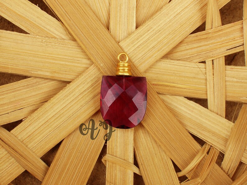 May include: A deep red faceted glass pendant with a gold-toned metal bail. The pendant is on a woven straw background.
