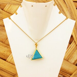 May include: A gold-colored necklace with a triangular pendant. The pendant features a vibrant turquoise stone set within a gold frame. The necklace is displayed on a white jewelry stand against a textured, light brown background.