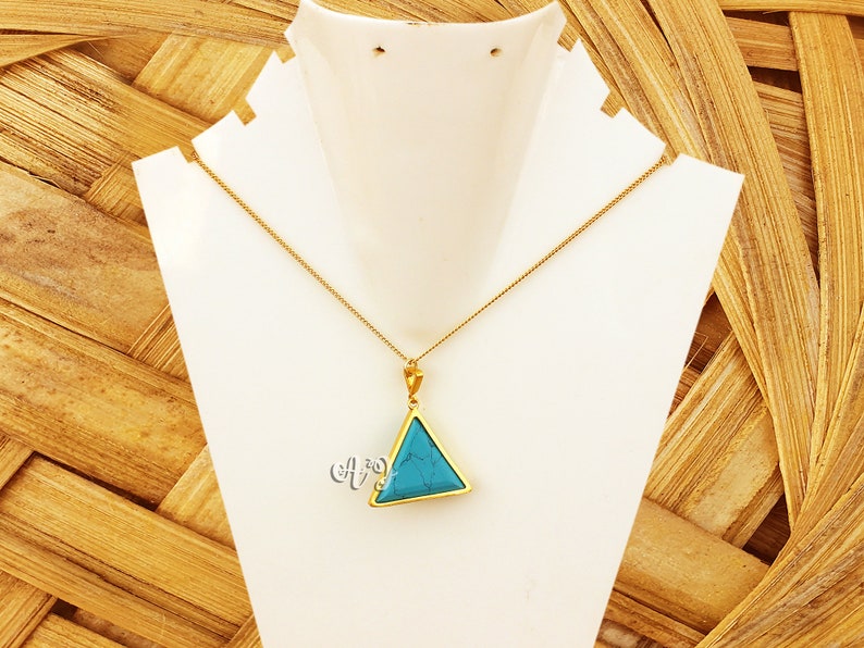 May include: A gold-colored necklace with a triangular pendant. The pendant features a vibrant turquoise stone set within a gold frame. The necklace is displayed on a white jewelry stand against a textured, light brown background.
