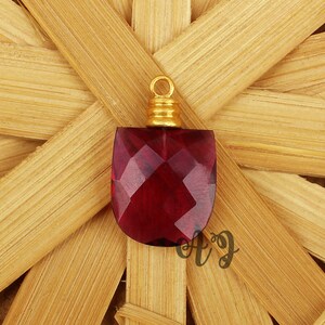 May include: A red faceted gemstone pendant with a gold-toned bail. The pendant is on a woven bamboo background.
