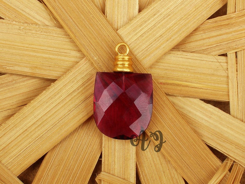 May include: A red faceted gemstone pendant with a gold-toned bail. The pendant is on a woven bamboo background.