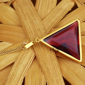 May include: A gold necklace with a triangular pendant featuring a red gemstone. The pendant is set in a gold frame.