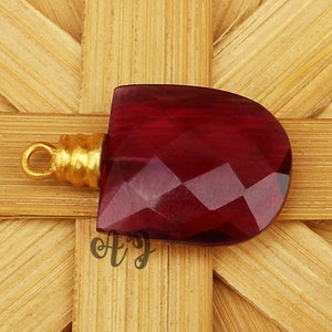 May include: A deep red faceted gemstone pendant with a gold-toned bail. The pendant is on a woven bamboo background.