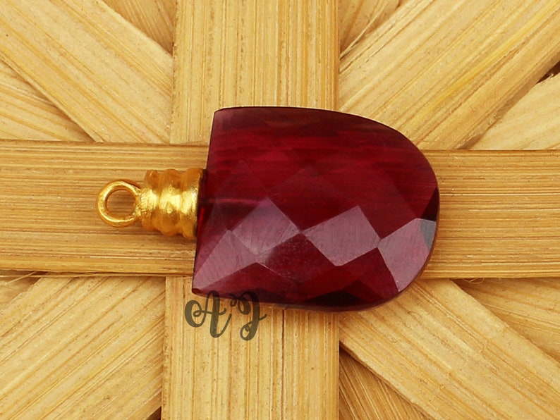 May include: A deep red faceted gemstone pendant with a gold-toned bail. The pendant is on a woven bamboo background.