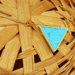 May include: A gold-colored necklace featuring a triangular pendant with a vibrant turquoise stone. The pendant is framed by a gold border and hangs from a delicate gold chain. The turquoise stone has a unique, natural pattern.